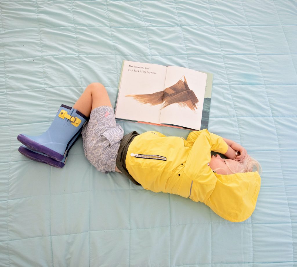 Additional and more specific support. Photo of a child in a coat and wellies laying on a bed reading a book.